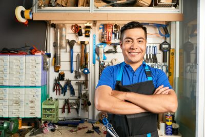 Portrait of happy confident mechanic in apron standing at workbench with various tools hanging behind him